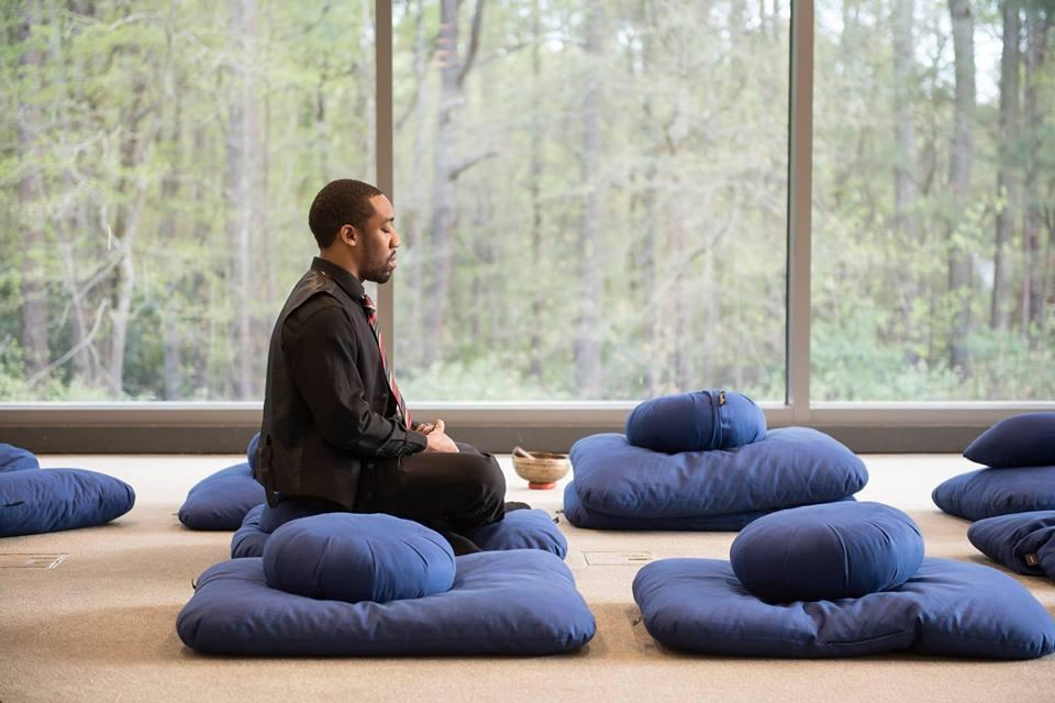 Black man sitting on top of two meditation pillows inside of a glass room that is surrounded by nature. Black man sitting on top of two meditation pillows inside of a glass room that is surrounded by nature.